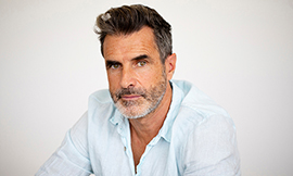 headshot of a man with dark hair, a dark and gray beard and wearing a light blue collared shirt and smiling at the camera